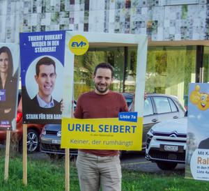 Poltiker "Uriel Seibert", der mit einem Werbeplakat von sich posiert